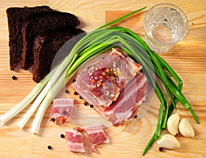 A piece of lard, a glass of vodka, garlic, green onions, peas and slices of rye bread are on a wooden stand