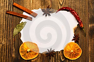 Piece of paper on wooden background. Spices, kitchen herbs lay around white paper. Cinnamon sticks, dried orange and