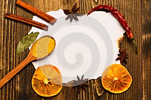 Piece of paper on wooden background. Spices, kitchen herbs lay around white paper. Cinnamon sticks, dried orange and
