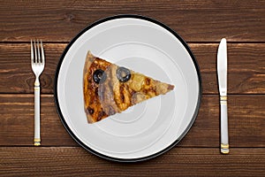 A piece of pizza on white plate with a knife and fork on wooden background. Slice of tasty pizza on the plate. Top view