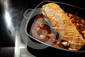 Piece of pork meat with incised fat rind on a brown sauce base from vegetables and red wine in a black cooking pan on the stove