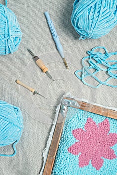 A piece of punch needle embriodery handiwork in a frame, three punch needles and three henks of blue velour yarn on linnen fabric
