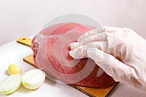 A piece of raw pork on a chopping board, pressed by a gloved hand. Next to it are onions and garlic. Cooking