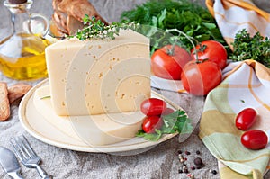 A piece of semi-hard cheese on a beige plate. Garnished with cherry tomatoes, parsley and a branch of temen. In the background is