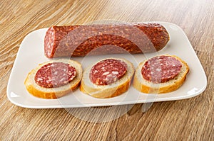 Piece of smoked sausage, sandwiches in plate on wooden table