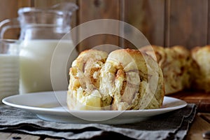 A piece of yeast cake with cinnamon on a white plate in the foreground and a glass jug of milk in the background on a wooden table