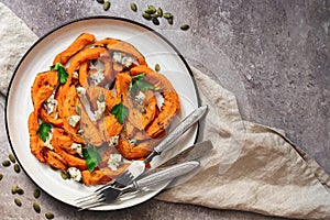 Pieces of baked pumpkin with herbs, seeds and cheese in a plate on a beige textured background. Top view, flat lay