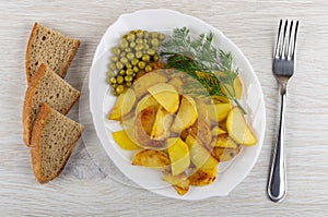 Pieces of bread, plate with fried potatoes, green peas and dill, fork on table. Top view