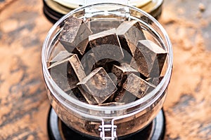 Pieces of dark bitter chocolate with cocoa in a glass jar on wooden background. Concept of confectionery ingredients