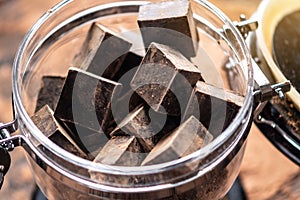 Pieces of dark bitter chocolate with cocoa in a glass jar on wooden background. Concept of confectionery ingredients
