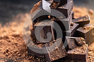 Pieces of dark bitter chocolate with cocoa powder on dark wooden background. Concept of confectionery ingredients