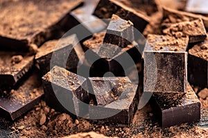 Pieces of dark bitter chocolate with cocoa powder on dark wooden background. Concept of confectionery ingredients