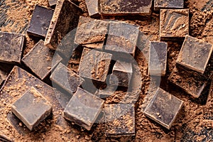 Pieces of dark bitter chocolate with cocoa powder on dark wooden background. Concept of confectionery ingredients