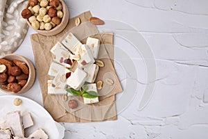 Pieces of delicious nutty nougat, hazelnuts and almonds on white textured table, flat lay. Space for text