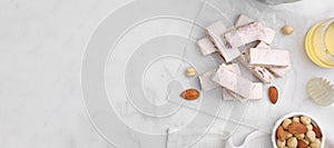 Pieces of delicious nutty nougat and ingredients on white marble table, flat lay. Banner design with space for text