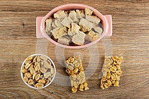 Pieces of halva in oval bowl, peanuts in white bowl, pieces of kozinak on table. Top view