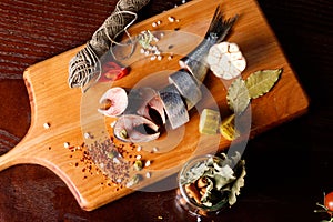 pieces of herring on a cutting Board, next to pepper, garlic, tomatoes on a branch ,parsley, pickled cucumber.