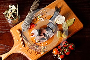 pieces of herring on a cutting Board, next to pepper, garlic, tomatoes on a branch ,parsley, pickled cucumber.