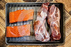 Pieces of pork and pieces of salmon on rack in broiling pan ready to cook sitting on rustic wood surface