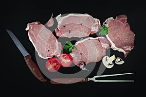 Pieces of raw roast beef meat for grilling on dark background with parsley, garlic, champignons, tomato and meat knife