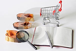 Pieces of red smoked salmon, magnifying glass, supermarket trolley and notepad with pen. Pre-sale preparation of fish. Quality