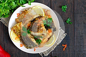 Pieces of roasted carp with lemon and greens on a plate on a dark wooden background.