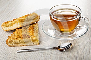 Pieces of shortbread pie with jam, cup with tea on saucer, spoon on wooden table