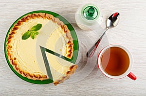Pieces of sour cream pie with spearmint in plate, sugar bowl, spoon, cup of tea on wooden table. Top view