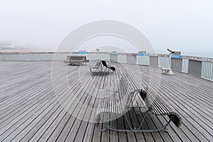 The Pier of Hastings on a misty summer day with three benches and telescopes, no people, sea and buildings in the distance