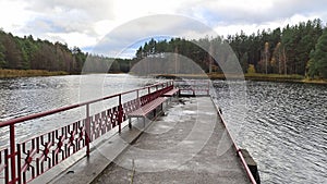 A pier with metal railings is built on the water near the lake shore. On the water ripples. There are reeds in the water. On the o