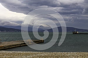 Pier on the sea beach and the lighthouse on the background of mountains and overcast sky.