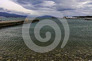 Pier on the sea beach and the lighthouse on the background of mountains and overcast sky.