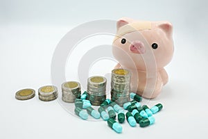 A piggy bank with a bandage at head behind stacking coins and blue green capsules pillon white background
