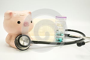 A piggy bank with a bandage at head and a stethoscope at chest with pill container and soft focus book bank