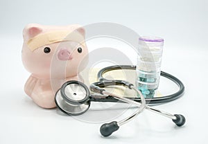 A piggy bank with a bandage at head and a stethoscope at chest with pill container and soft focus book bank on white background ,