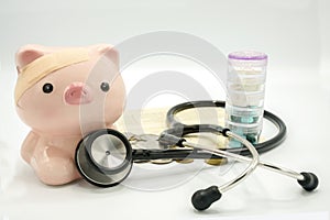 A piggy bank with a bandage at head and a stethoscope at chest with pill container and soft focus book bank on white background
