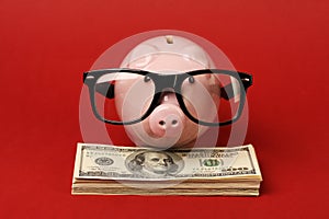 Piggy bank with black spectacle frame of glasses standing on stack of money american hundred dollar bills on red background