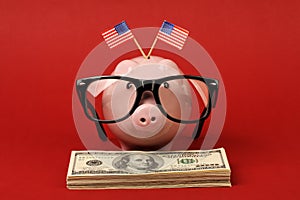 Piggy bank with black spectacle frame of glasses and two small USA flags standing on stack of money american hundred dollar bills