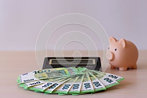 A piggy bank, a calculator and paper money in denominations of 100 euros, spread out in a fan on the table. The concept of