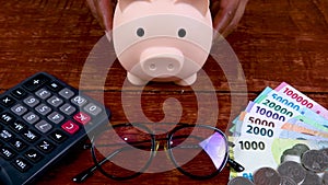 Piggy bank with glasses, calculator, and stacks of money, indonesian currency on wooden background