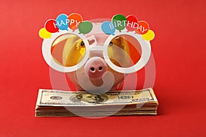 Piggy bank with Happy birthday party glasses standing on stack of money american hundred dollar bills on red background