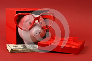 Piggy bank in love with red heart sunglasses standing in gift box with ribbon and with stack of money american hundred dollar bill