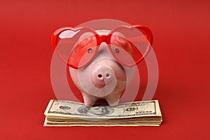 Piggy bank in love with red heart sunglasses standing on stack of money american hundred dollar bills on red background