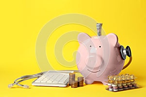 Piggy bank with money, stethoscope, calculator and pills on yellow background, space for text. Medical insurance