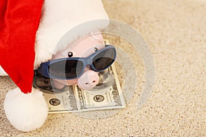 Piggy bank with Santa Claus hat standing on towel from greenback hundred dollars with sunglasses on the beach sand