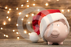 Piggy bank with Santa hat on table against blurred Christmas lights.