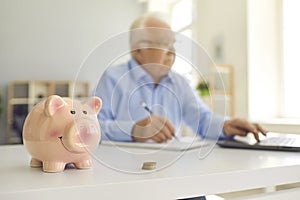 Piggy bank standing on desk with blurred retired man using laptop and doing accounting in background