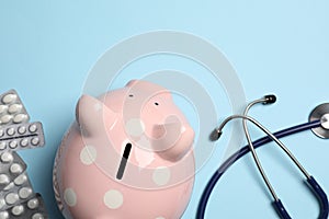 Piggy bank, stethoscope and pills on light blue background, flat lay. Medical insurance
