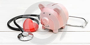 Piggy bank with stethoscope and red heart on white wooden table