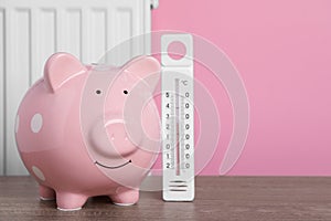 Piggy bank with thermometer on wooden table near heating radiator. Space for text